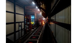 Innenansicht einer Entstaubungsanlage. Im Vordergrund links sieht man die Außenwand der Einhausung mit Gitterstrukturen und eine offene Tür. Rechts im Bild ist ein langer Tunnel sichtbar, in dem die Gießstraße installiert ist. Am Ende des Tunnels sind Lichter und Arbeiter zu erkennen. Der Fokus liegt auf einem beleuchteten Bereich in der Mitte des Tunnels. | © INFRANORM®